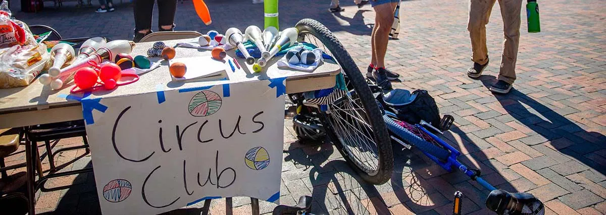 A student juggles behind a table with a handwritten sign reading "Circus Club" as two people stand nearby chatting