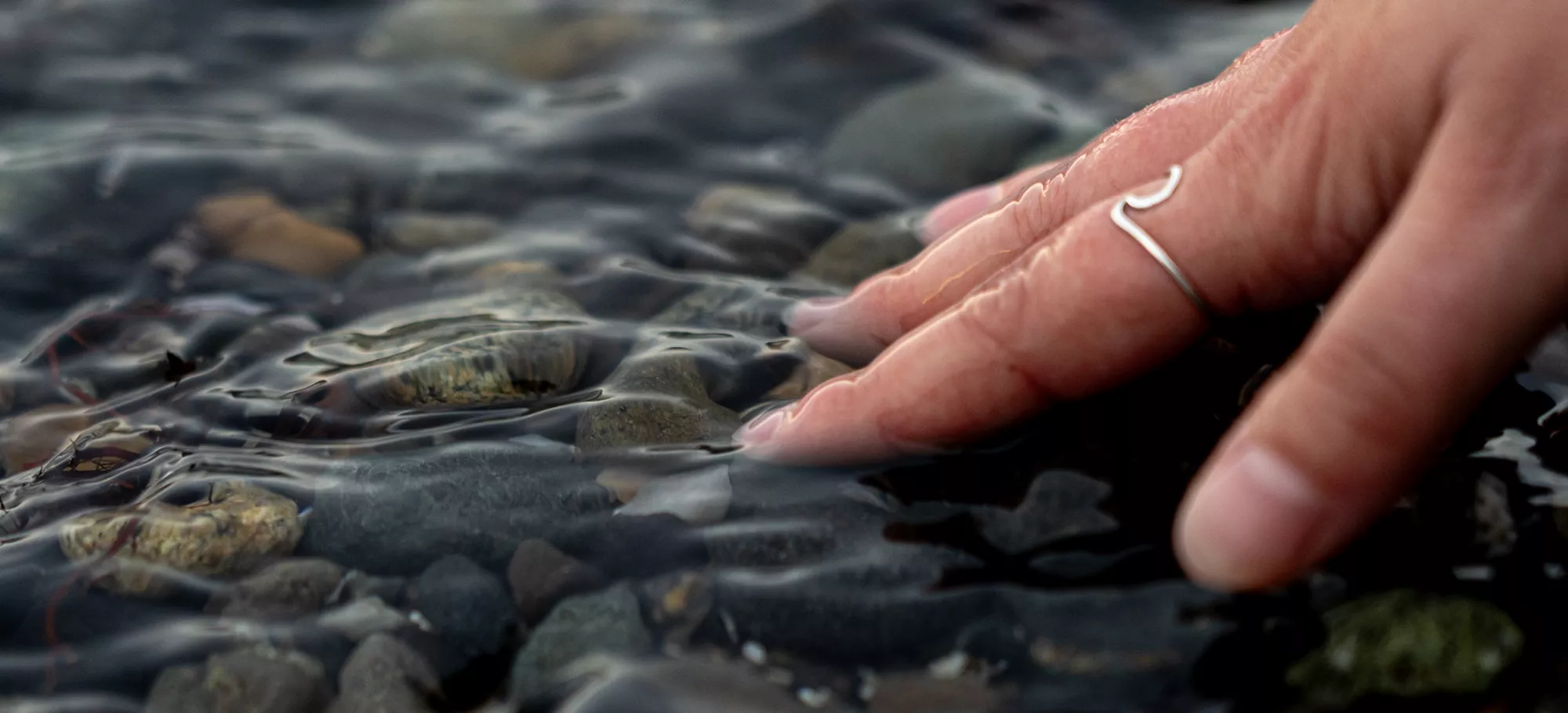 A hand with a ring gently touching the water's surface, creating ripples.