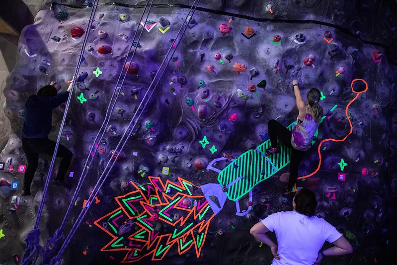 Student climbing a rock wall with a black light glowing rocket painted on it