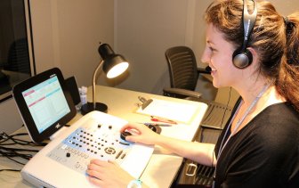 Graduate student on right with arms extended to another person's ear, fitting for hearing equipment