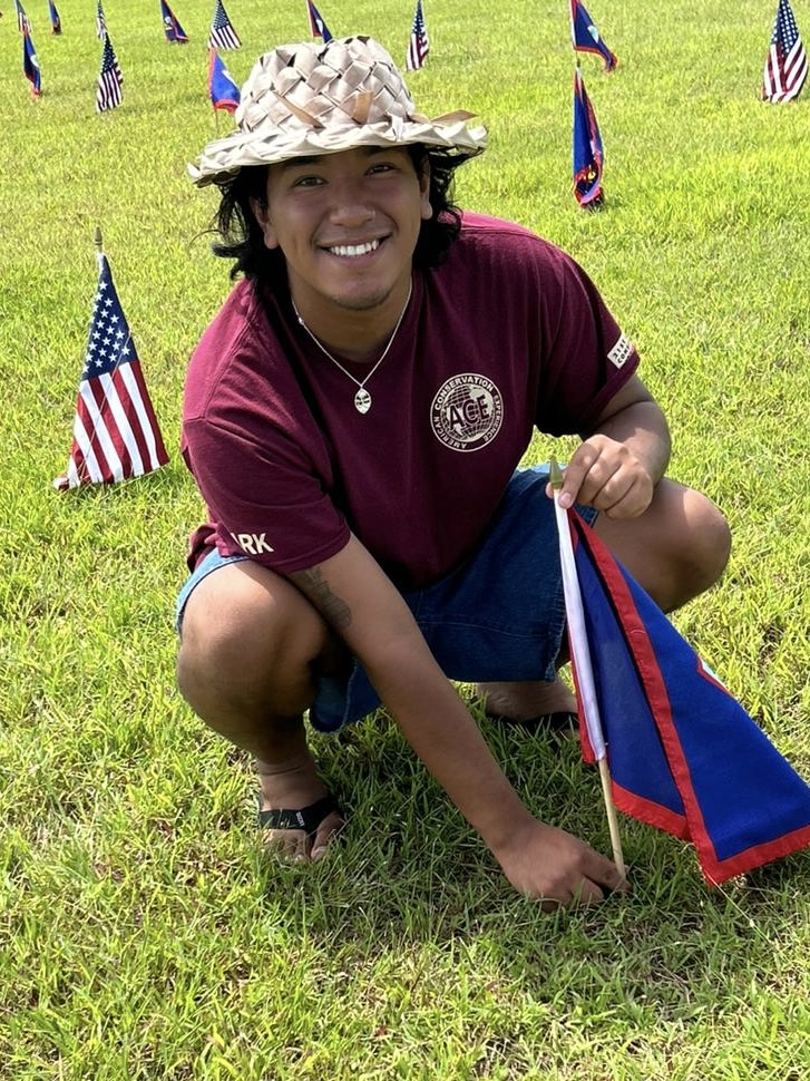 Favio Guzman-Estrada wearing a woven reed hat, squatting on the ground and placing a small Flag of Guam