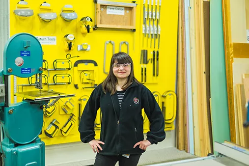 A student standing in front of a bright yellow tool wall, wearing safety glasses with hands on hips