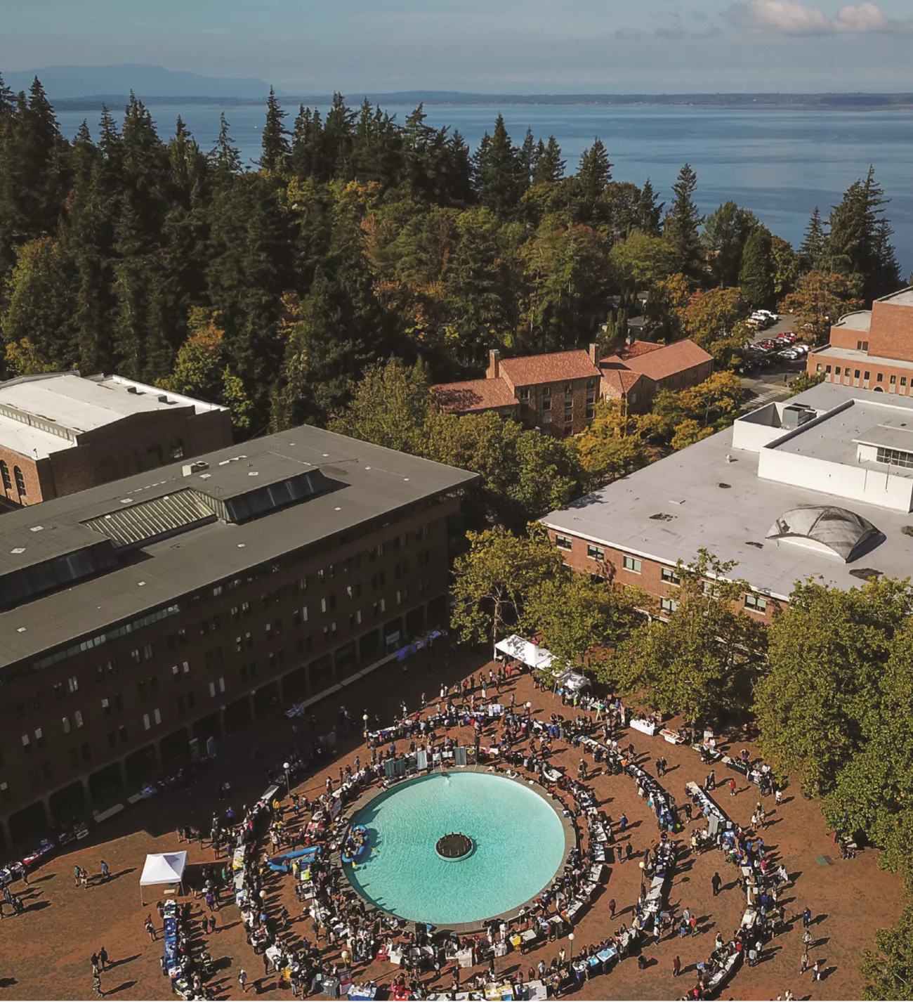Aerial view of a bustling university courtyard with a fountain, surrounded by trees and overlooking a large body of water.